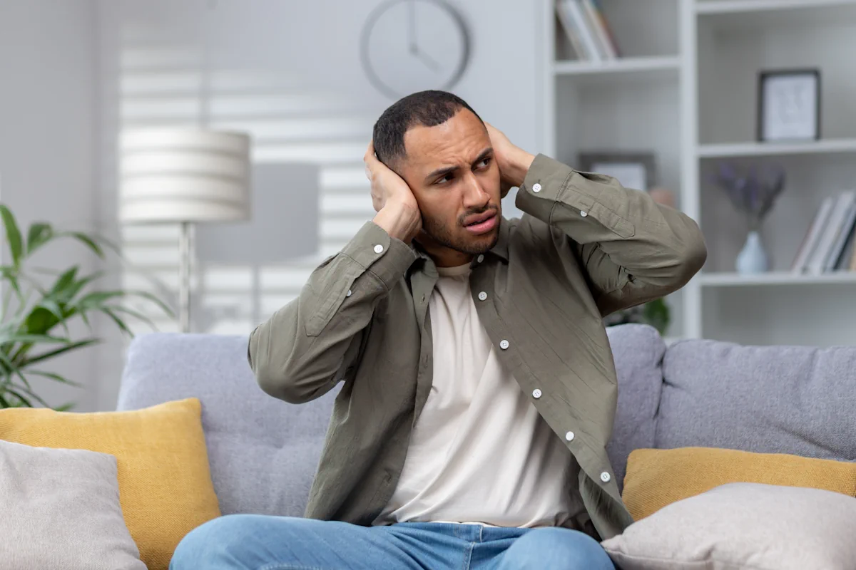 un homme assis dans son canapé qui se bouche les oreilles avec les mains